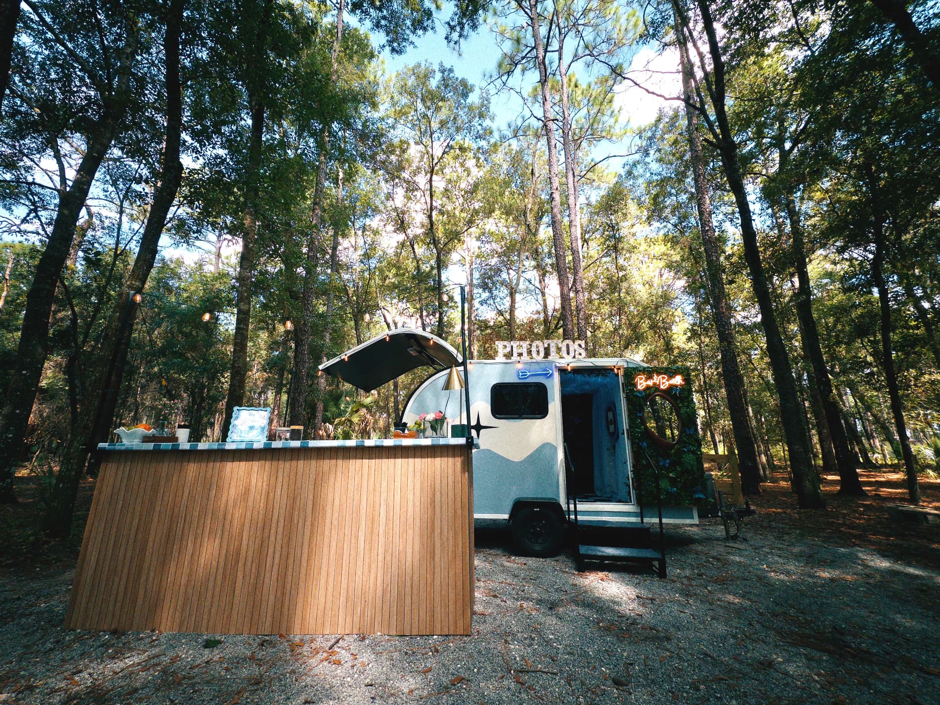 The Something Blue camper and L-shaped luxury bar set up in a wooded outdoor venue